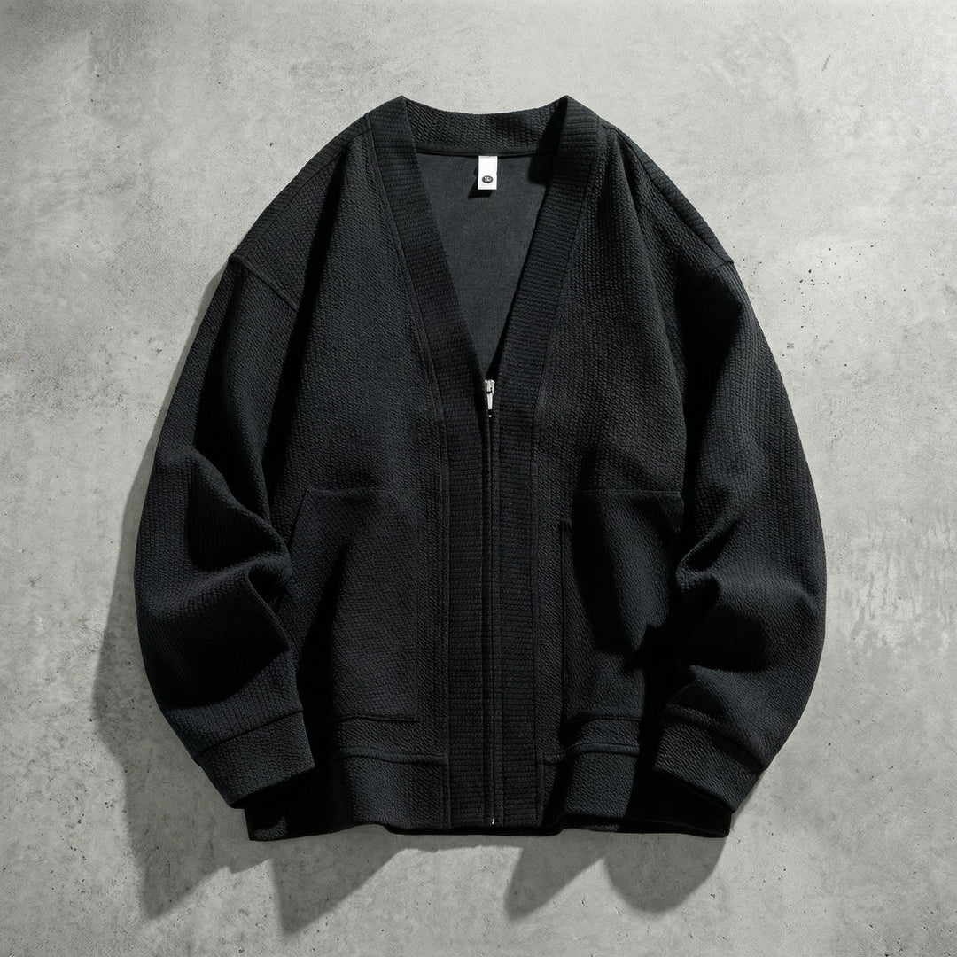 Cardigan zippé noir à col V posé à plat sur du béton, maille gaufrée et poches plaquées