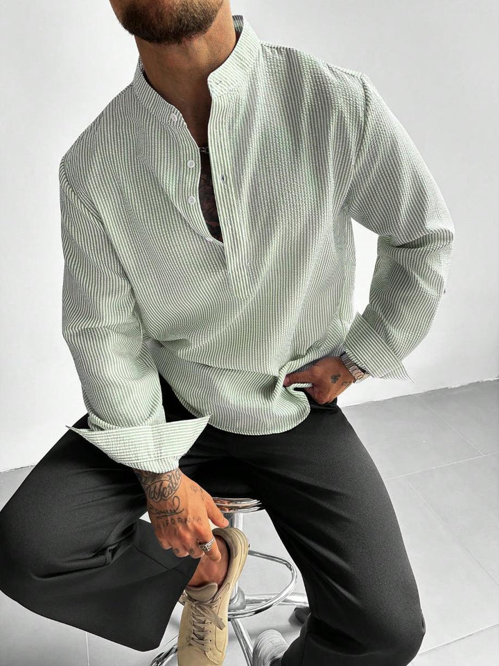 Homme assis sur un tabouret en chemise à fines rayures vert d'eau, col mao et pantalon noir