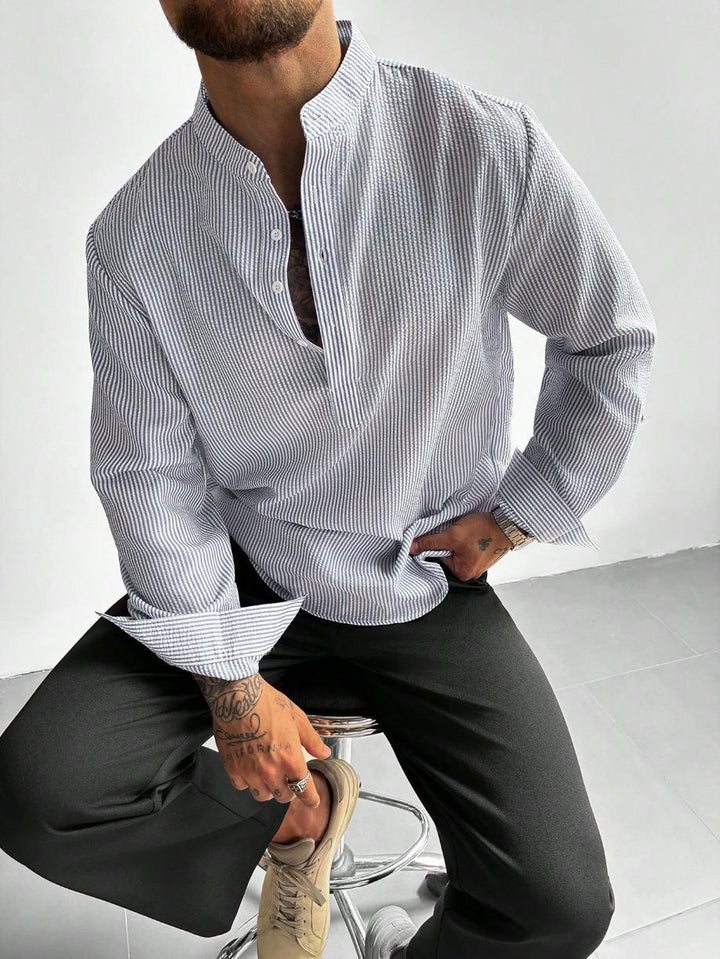 Homme assis en chemise à fines rayures grises, col mao ouvert et pantalon noir
