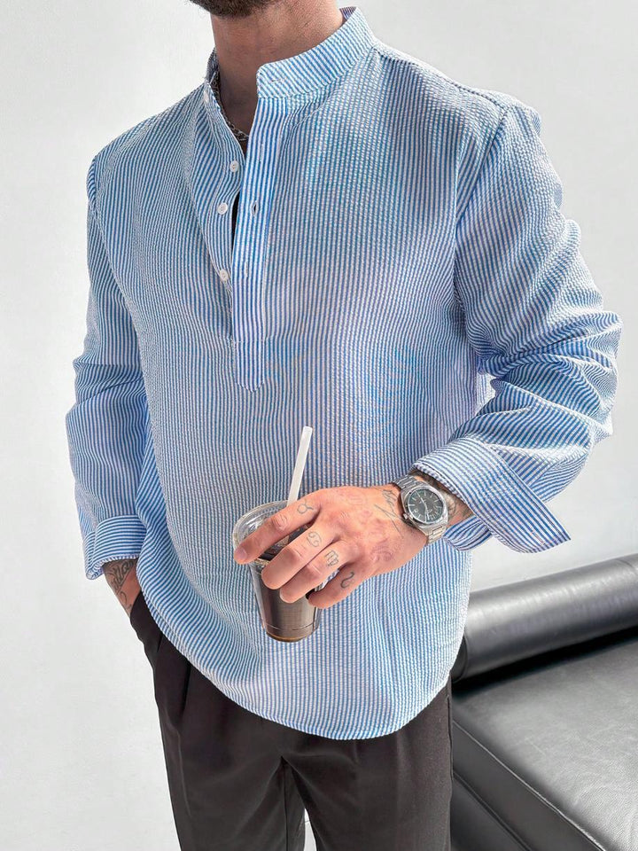 Chemise à rayures bleu ciel à col mao, portée avec un pantalon foncé, verre à la main