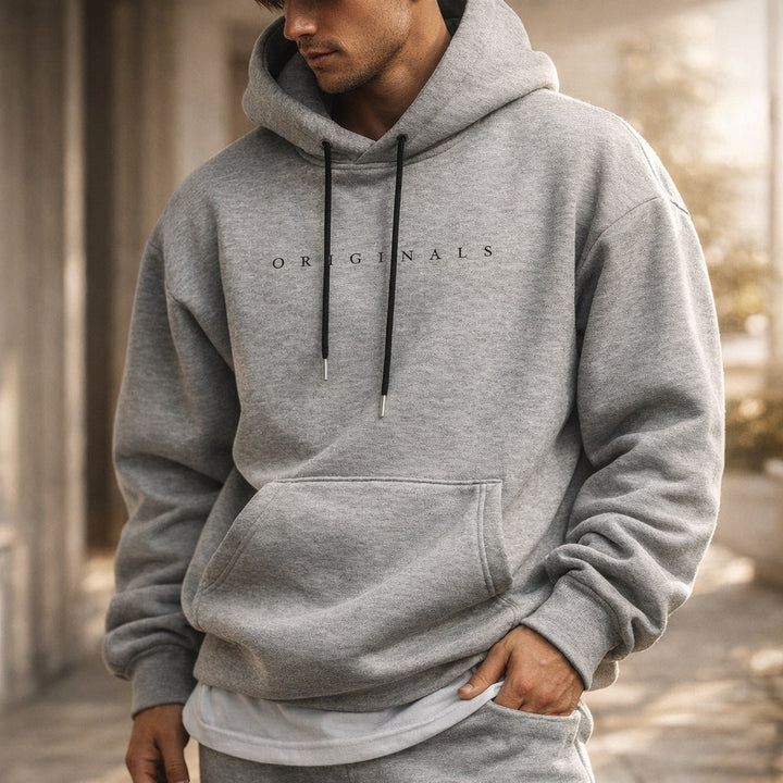 Homme en sweat à capuche gris chiné, capuche relevée, dans une rue ensoleillée