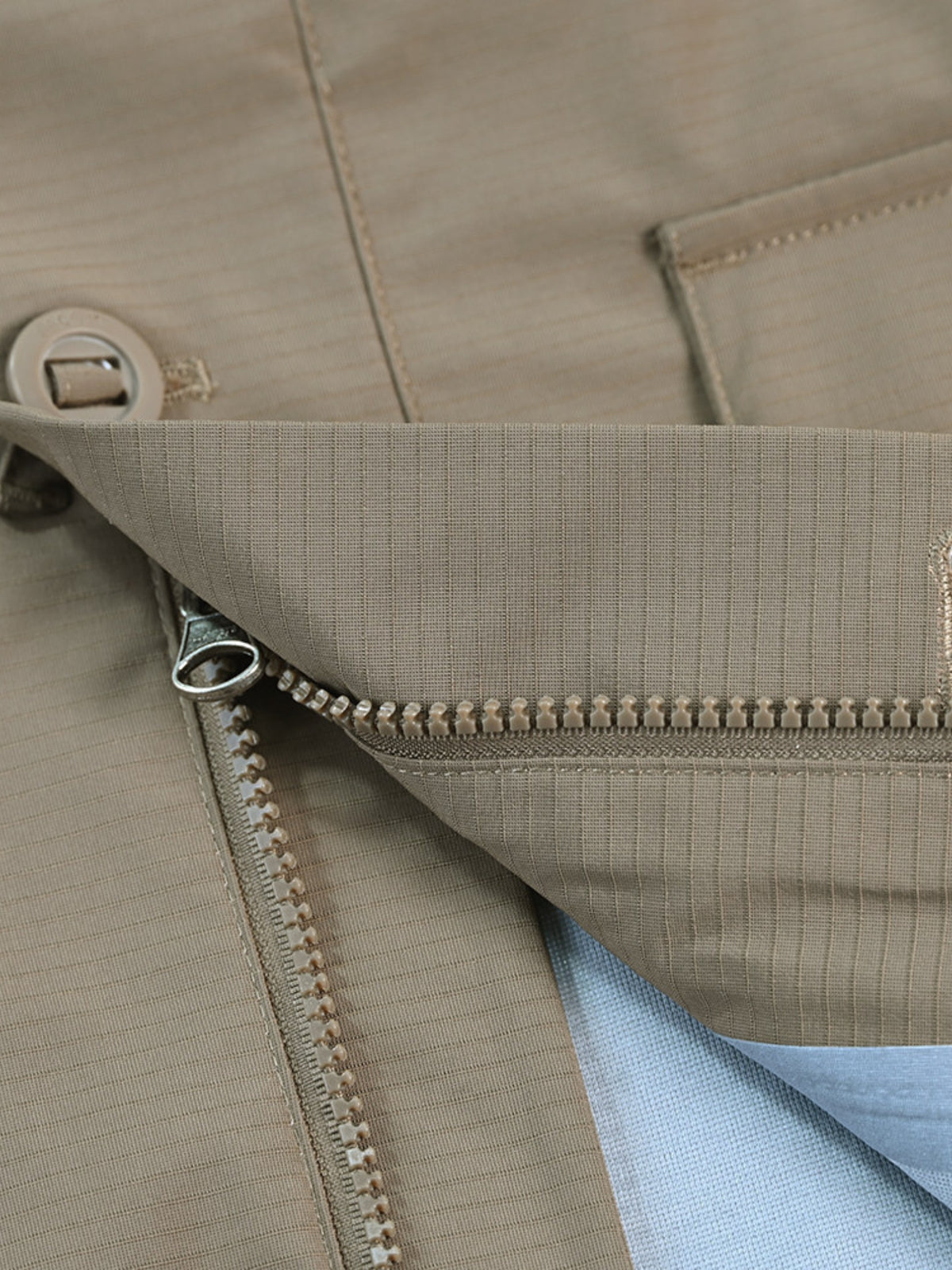 Gros plan sur le bas d'une veste beige, fermeture eclair et bouton pression, doublure bleu clair